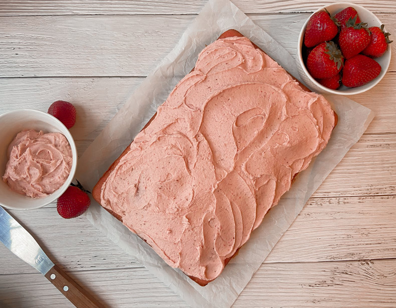 Une vue aérienne montre un gâteau rectangulaire avec du glaçage rose, reposant sur un plateau placé sur une surface de bois. Un petit bol de glaçage, un petit bol de fraises et une spatule à glaçage sont visibles près du plateau.