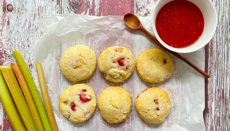 Six scones reposant sur un morceau de papier ciré posé sur une surface de bois usée. On voit des branches de rhubarbe fraîche, un bol de confiture rouge et une cuillère de bois.