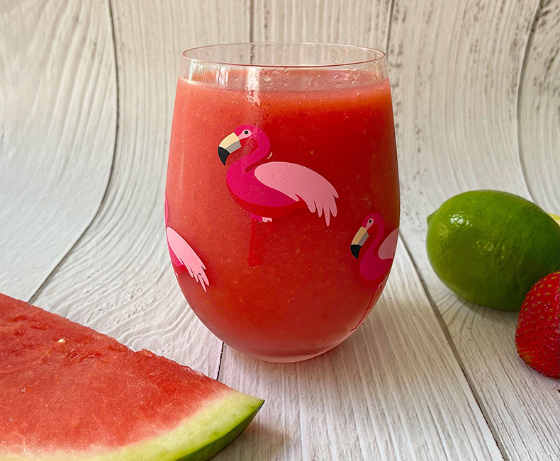 A clear glass decorated with flamingos is filled with watermelon slush. A slice of fresh watermelon and a lime sit next to the glass, against a wooden surface.