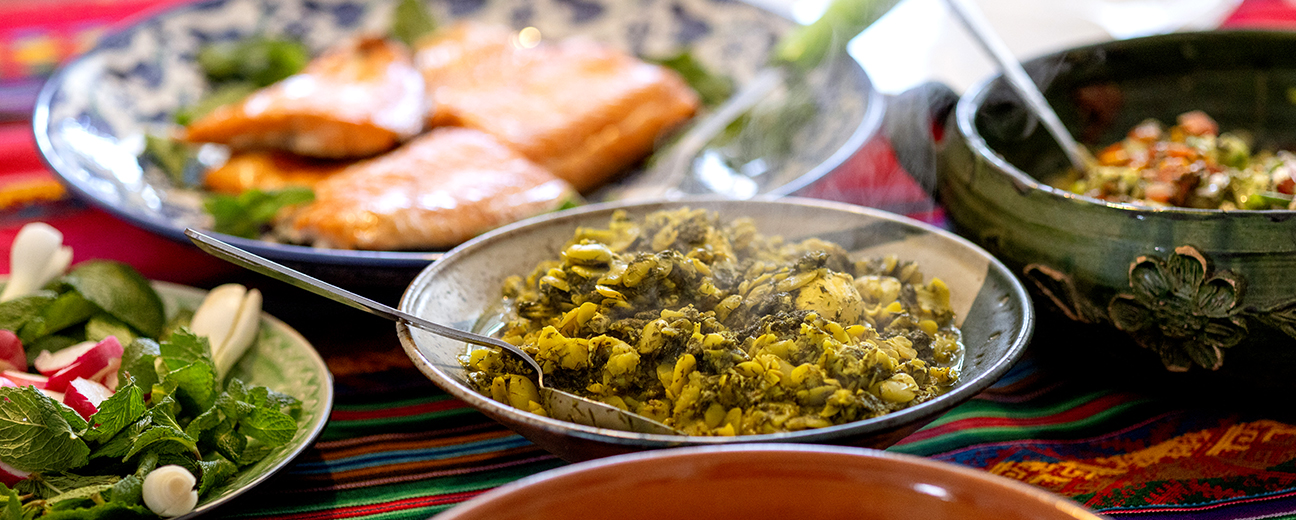 A vibrant spread of dishes on a colorful tablecloth, featuring steaming vegetable curry in a bowl, fresh greens and radishes on a plate, baked salmon fillets on a ceramic platter, and a salad in a decorative bowl.