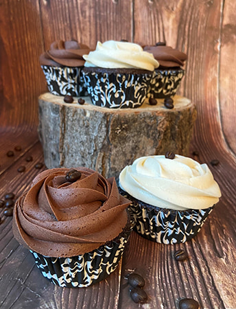 Five cupcakes in black-and-white liners are displayed on a wooden table, with a wooden background. Two cupcakes are topped with white icing and three are topped with brown icing.