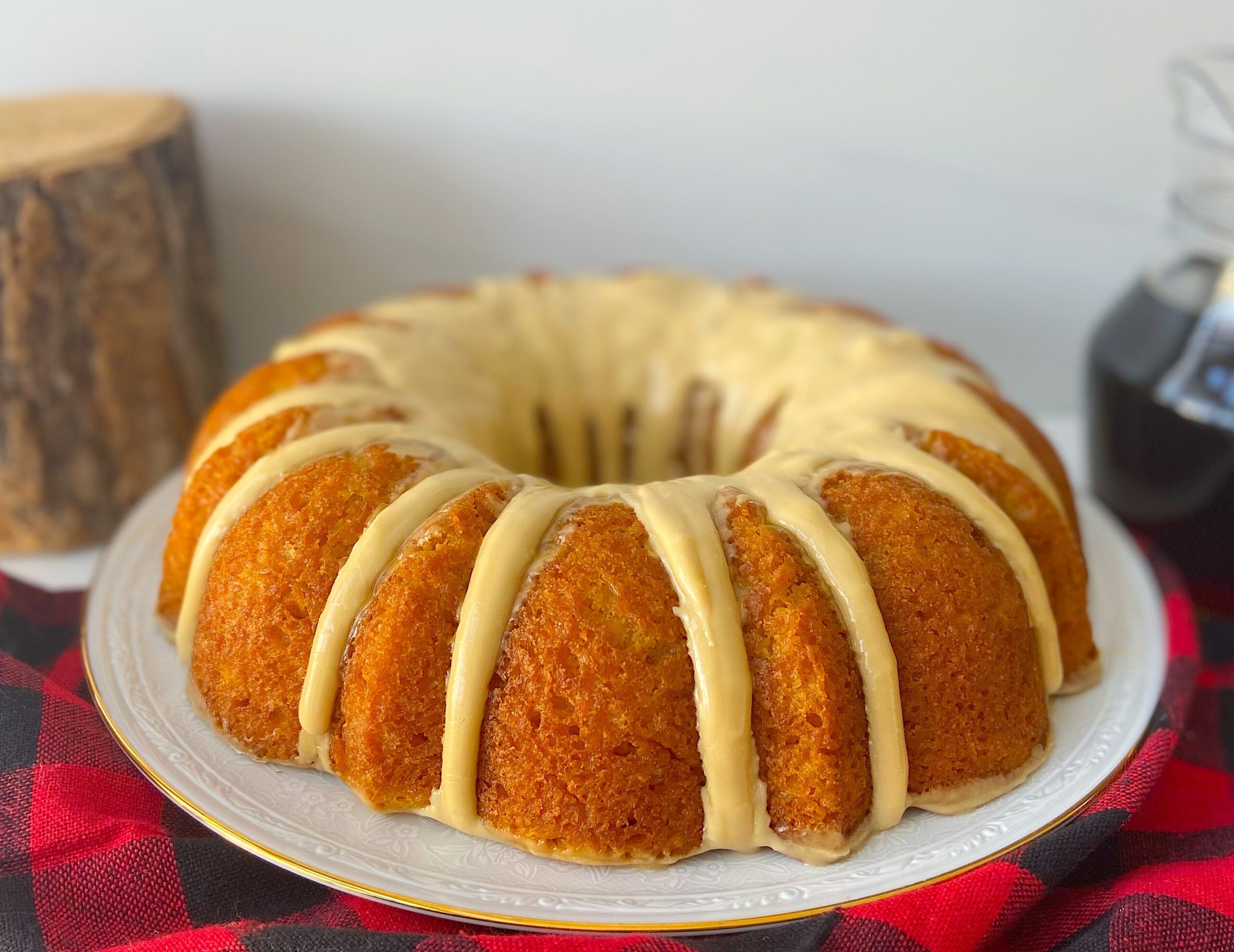 On peut voir un gâteau Bundt à l’érable sur une assiette blanche ronde qui repose sur une nappe à carreaux rouge et blanche. En arrière-plan, partiellement obscurcies, se trouvent une bouteille de sirop d’érable foncé et une bûche d’érable.