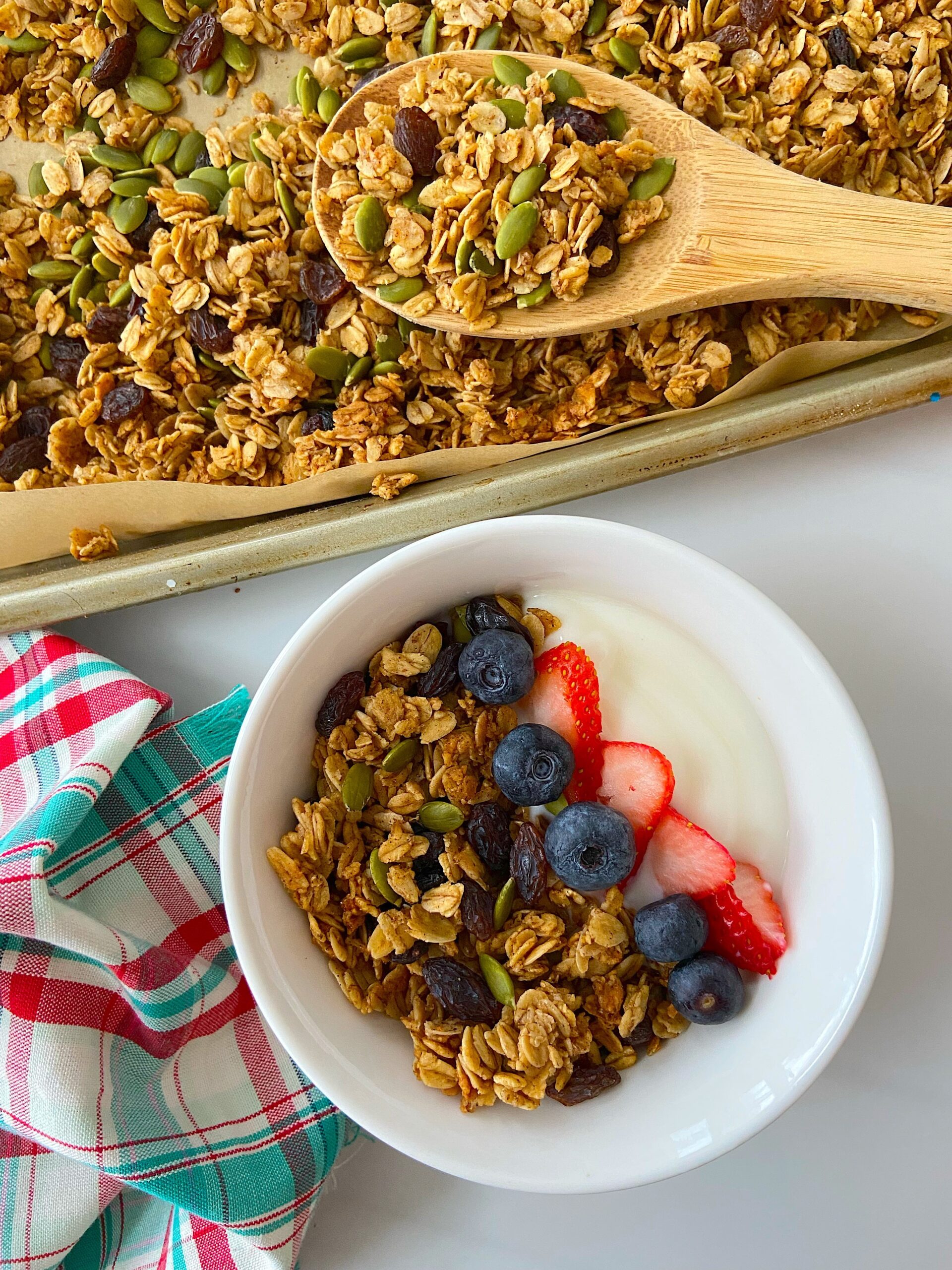 A bowl of granola, yogurt, and fresh fruit sits on a white surface. Next to it, a baking pan and a wooden spoon are filled with the freshly-baked granola.