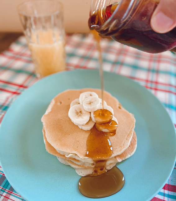 Quelqu’un verse du sirop d’érable à partir d’un pot en verre sur une pile de crêpes et des bananes tranchées. La nourriture repose sur une assiette bleue, placée sur une nappe à carreaux.