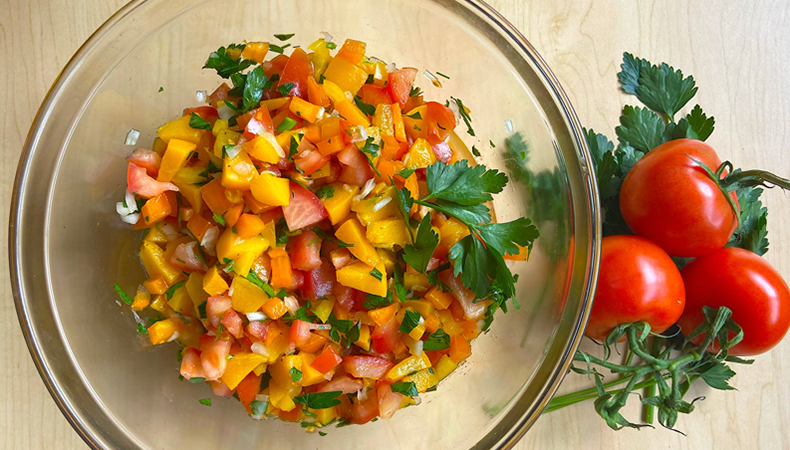 Une vue aérienne montre un bol transparent rempli de salsa fraîche reposant sur une table de couleur pâle. Trois tomates mûres sont placées près du bol.