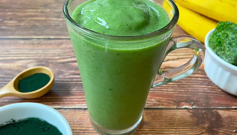 Un verre rempli d’une épaisse boisson verte repose sur une surface de bois rustique. Un bol et une cuillère avec de la poudre verte se trouvent à l'avant-plan, avec des épinards et des bananes à l'arrière-plan.