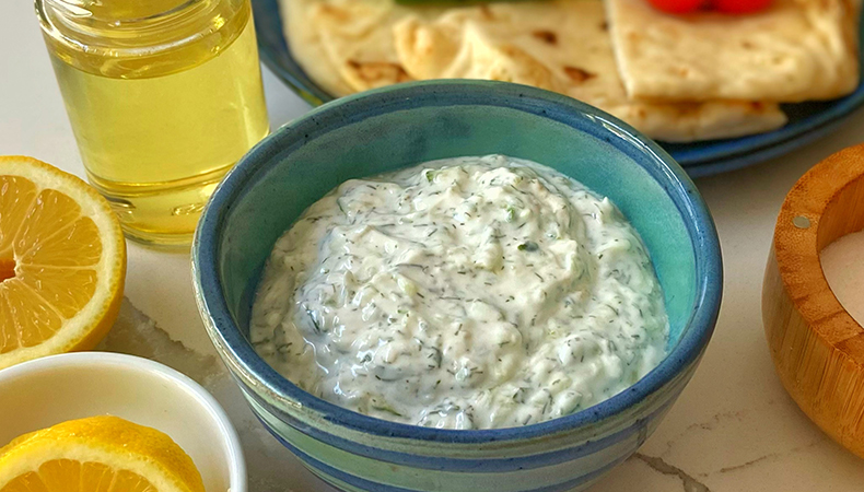 Un bol de sauce bleu est posé sur une table blanche. Des quartiers de citron, de l'huile d'olive et un bol de sel sont visibles autour du dip. Une assiette bleue contenant des légumes et des morceaux de pita est visible à l'arrière-plan.