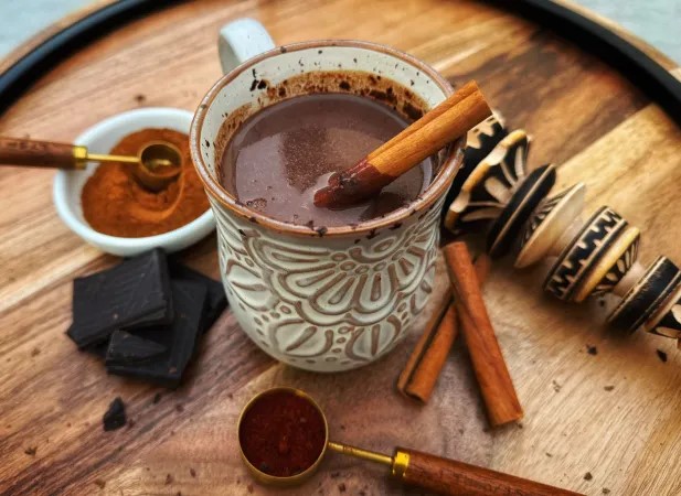 A mug of hot chocolate, garnished with a cinnamon stick, sits on a rustic surface. Nearby, a traditional molinillo whisk and an assortment of spices can be seen.