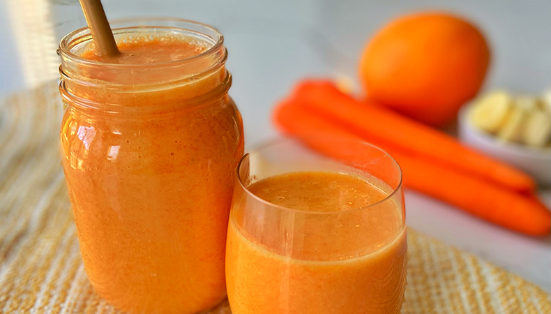 Two glasses filled with a thick, orange beverage sits on a yellow tablecloth on a white surface. An orange, carrots and banana slices are visible in the background.  