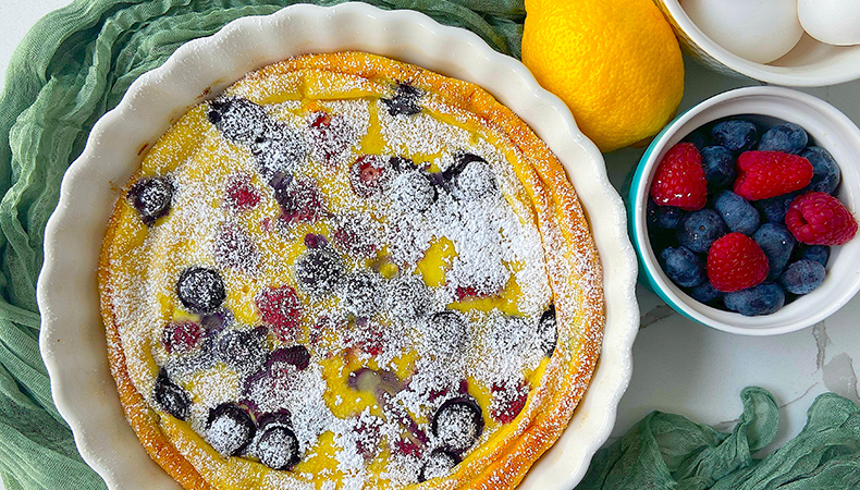 An aerial view shows a clafoutis sitting on a counter, surrounded by a decorative green cloth, bowls of berries and eggs, and a lemon.