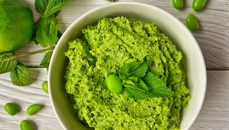 Une vue aérienne montre un bol de trempette verte sur une surface en bois clair. Des fèves d'edamame et des feuilles de menthe sont visibles autour de la base du bol.