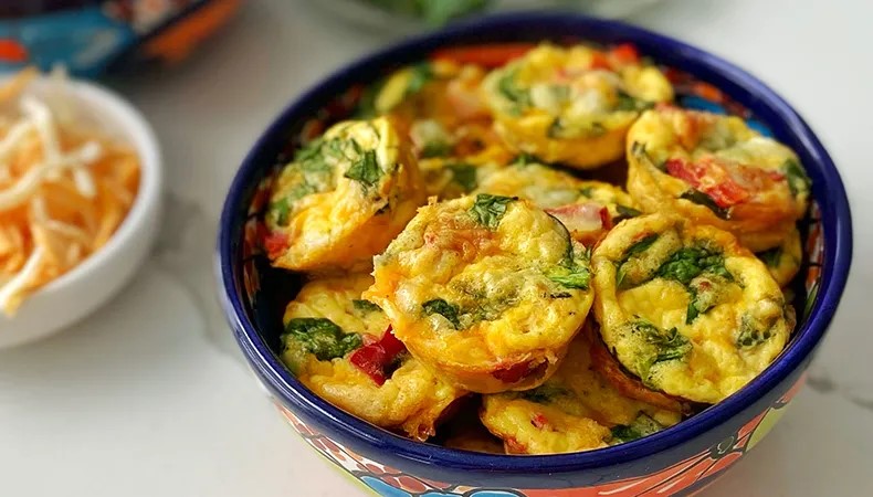 A bowl filled with egg bites sits on a white surface. Bowls of eggs, cheese, and vegetables are visible in the background.