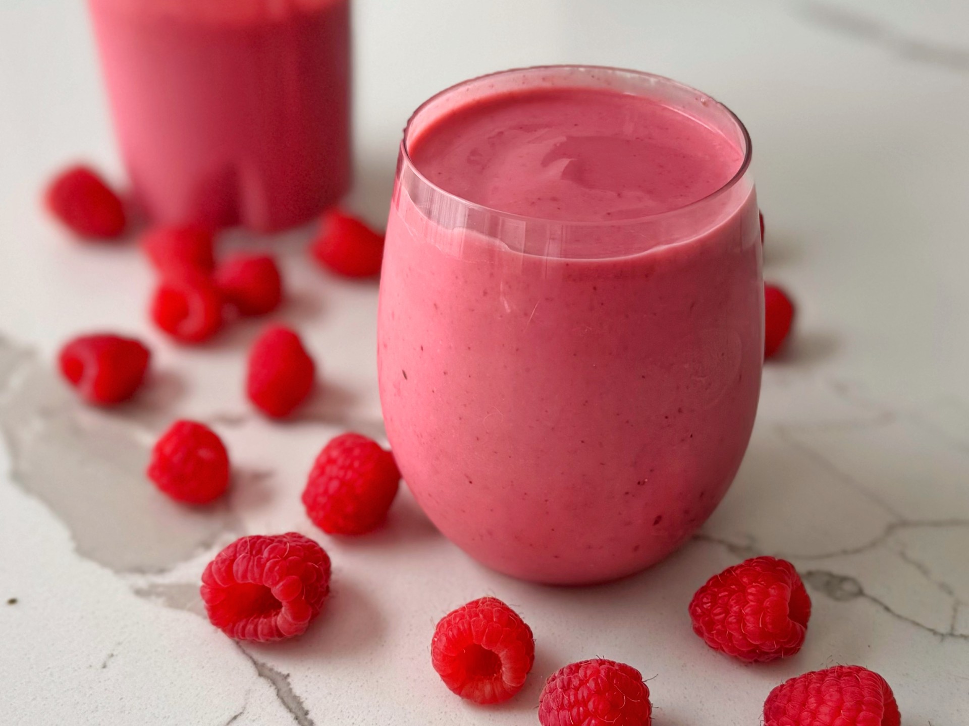 Un verre rempli d'une boisson rose épaisse est posé sur une surface blanche et grise. Une autre boisson rose se trouve en arrière-plan, avec des framboises fraîches éparpillées partout.