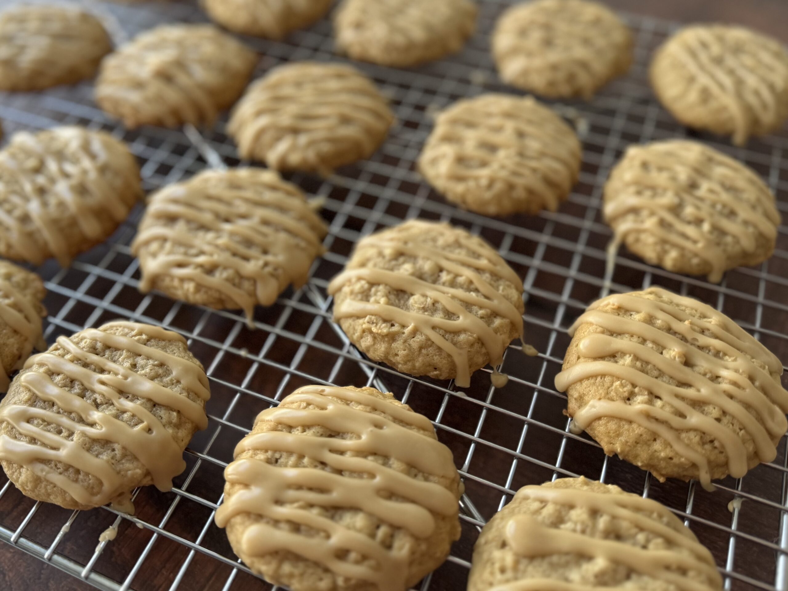 A stack of glazed maple cookies on a piece of parchment paper, with a cooling rack of more cookies in the background.