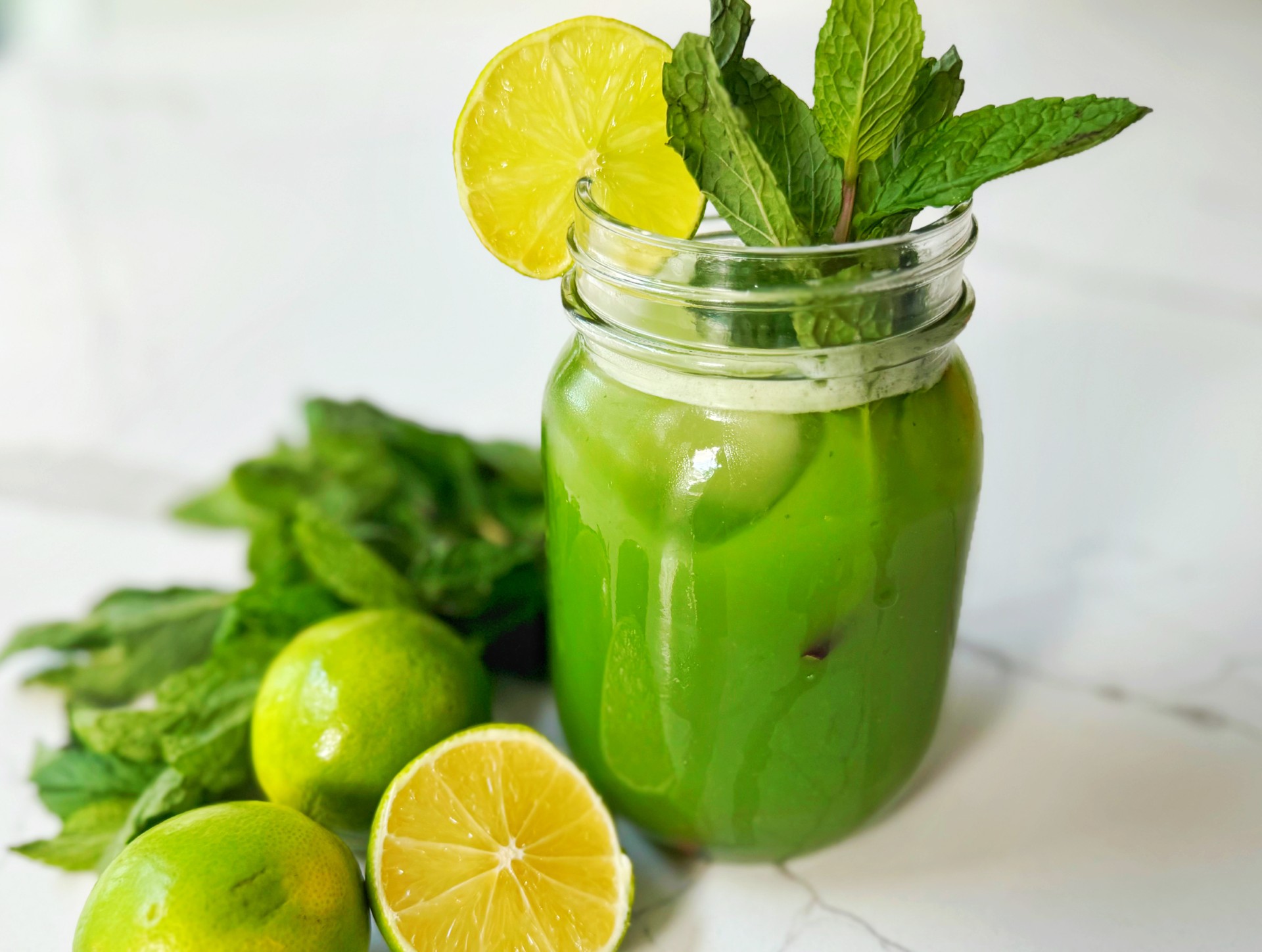 A mason jar filled with a creamy green beverage and garnished with lemon and mint. Limes and mint surround the jar