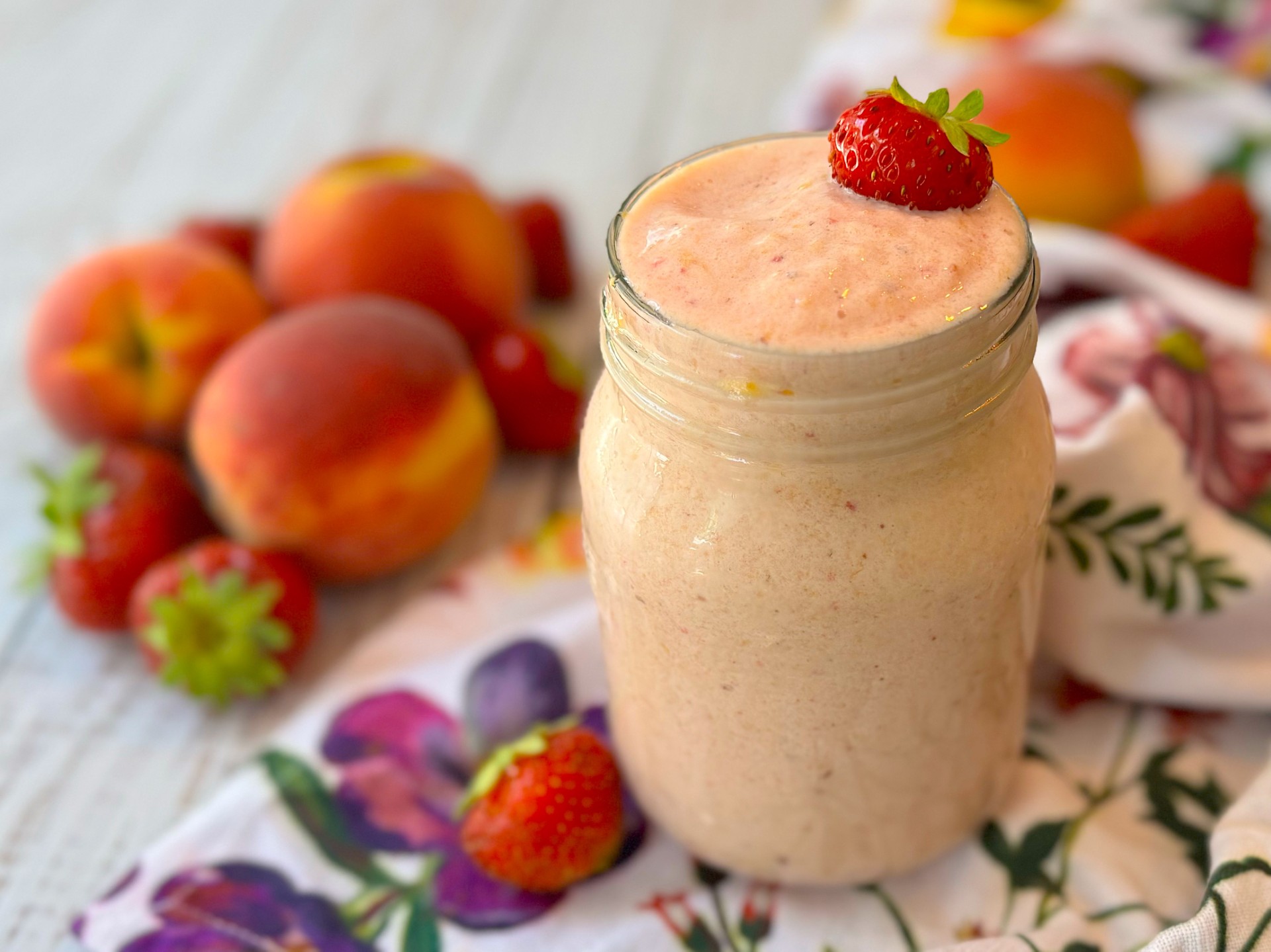 Glass jar filled with a creamy orange smoothie and topped with a strawberry. A variety of fresh fruit surrounds the glass