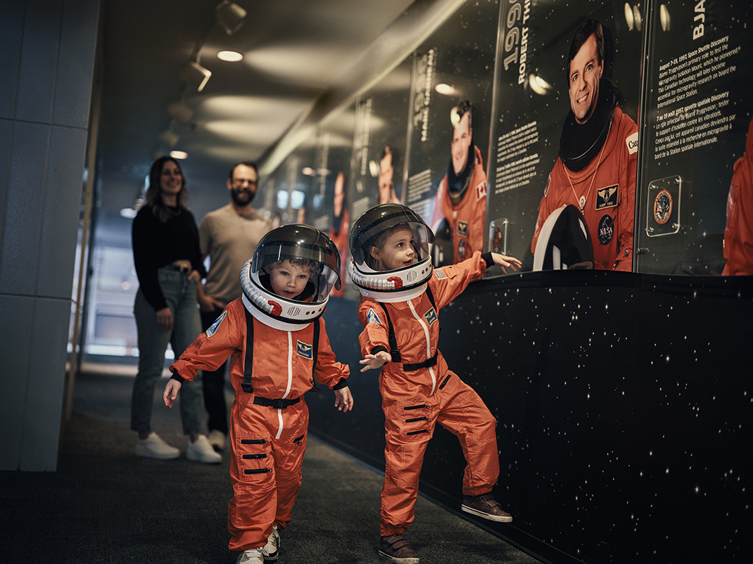 Two children in orange space suits walk next to a display of photos of Canadian astronauts.