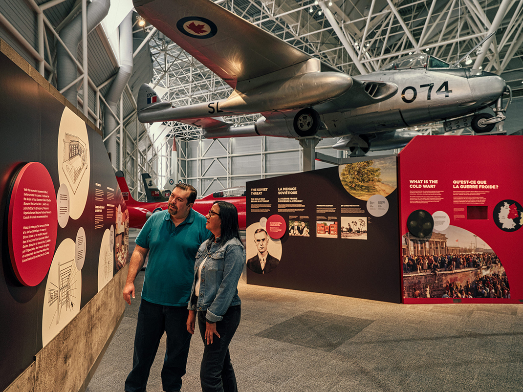 Two people look at an exhibition panel in the museum's Cold War section. In the background a silver aircraft is suspended over the exhibition.