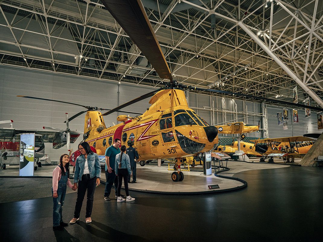 A museum exhibit displaying a large yellow Canadian military rescue helicopter. Visitors, including families, are gathered around, observing the helicopter and other aircraft in the background within a spacious hangar with a high, structured ceiling.