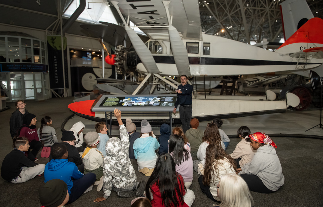 A group of students sits on the floor watching as a guide animates a lively discussion. The guide is standing in front of a white and black float plane inside the Canada Aviation and Space Museum.