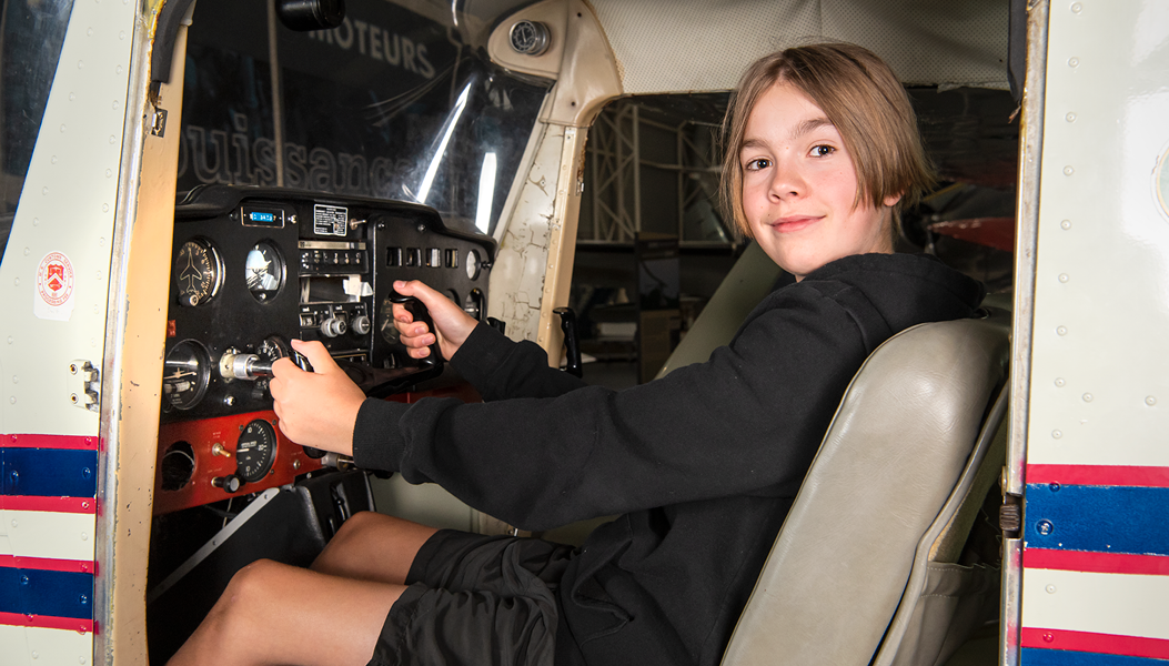 A blond boy with chin length hair and a black long sleeved top and black shorts sits at the controls of a Cessna aircraft.