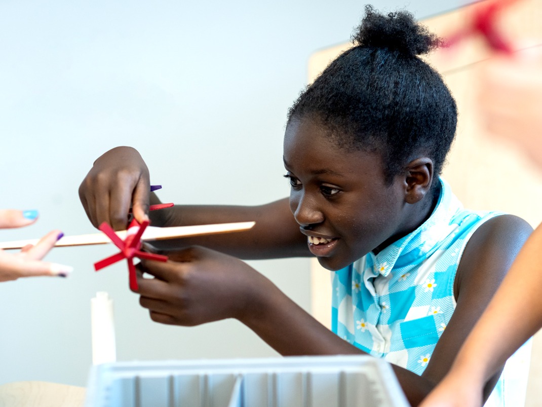 A young black girl with with dark hair in a bun and a blue and white shirt, is smiling as she holds up a handmade wooden airplane with a red propeller.