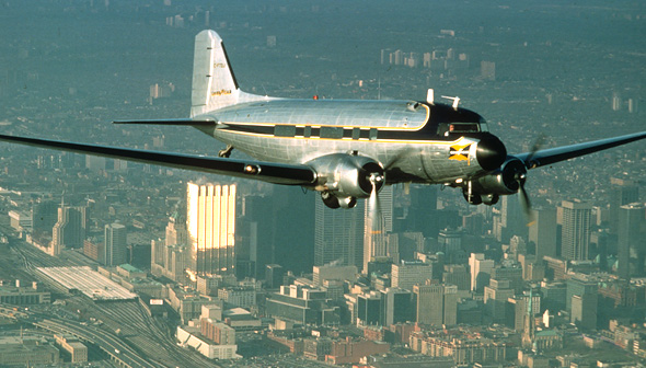 Silver DC-3 aircraft in flight over a city skyline