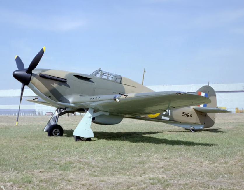 Un avion à hélices argenté sur l'herbe avec un ciel bleu derrière lui. L'avion est un Hawker Hurricane XII.