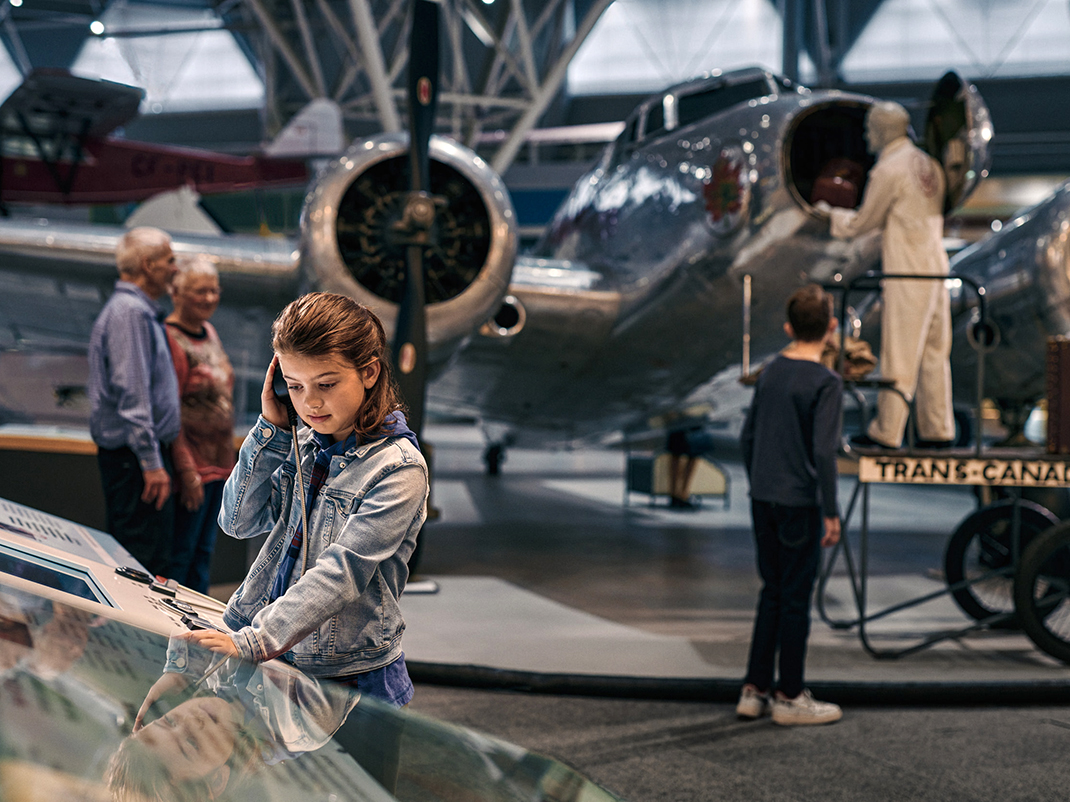 A young person listens to an audio recording at a museum exhibition. In the background other visitors tour aircraft on display