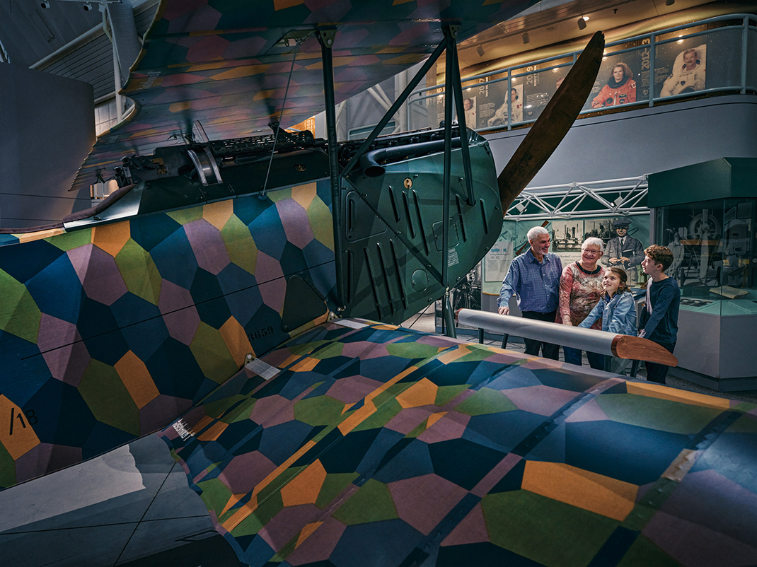 Museum visitors look at a Fokker aircraft with a yellow, pink, green and blue camouflage pattern.