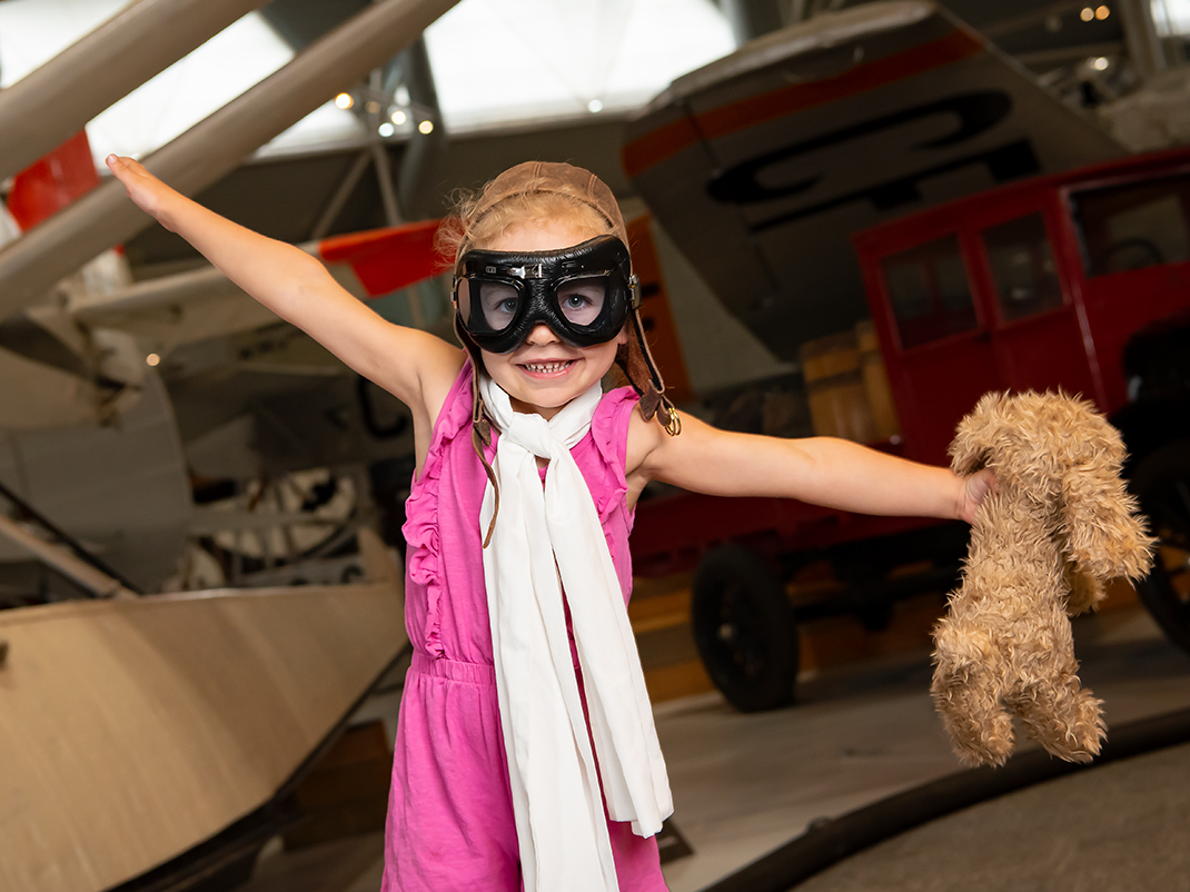 A young child holding a teddy bear and wearing aviator goggles holds her arms out like an airplane. There are historic aircraft in the background.