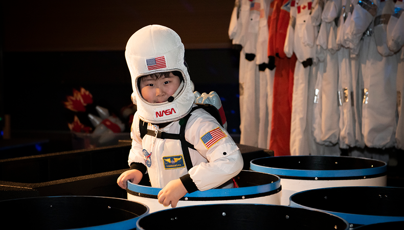 A small boy wears a space costume while sitting in one of several half cylinders on the museum stage.