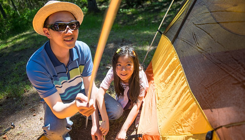 An adult and a child look up at the pole of a tent they are setting up.