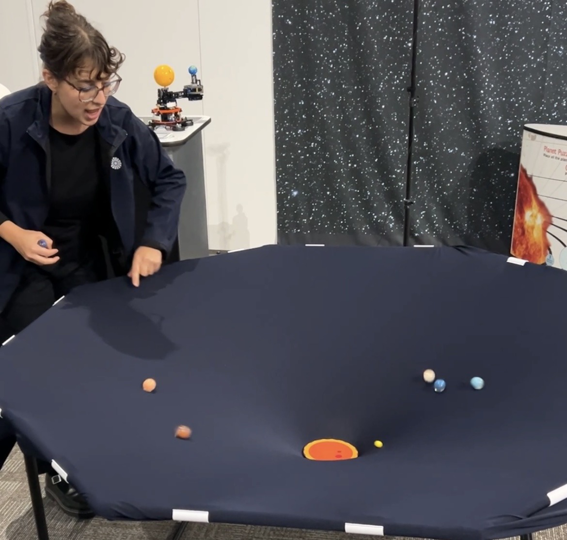 A person with dark hair in a bun is dressed in a navy jacket and has glasses on. The person is standing over a table with black stretchy cloth over it, at the centre there is a small sun and marbles are moving around it.