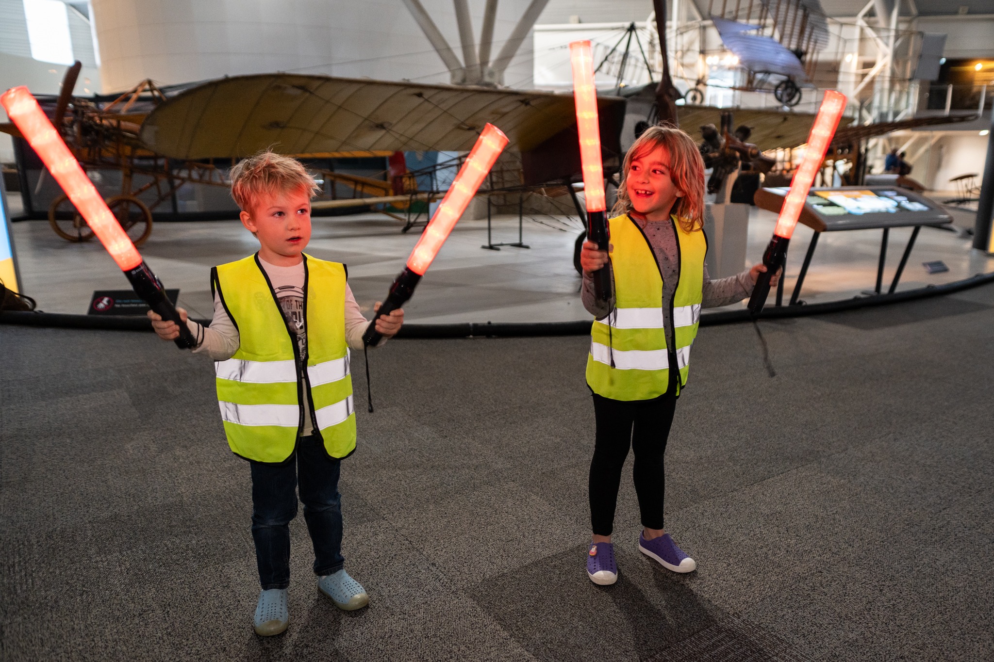 two small children wear vests and hold lit orange marshalling wands in the museum