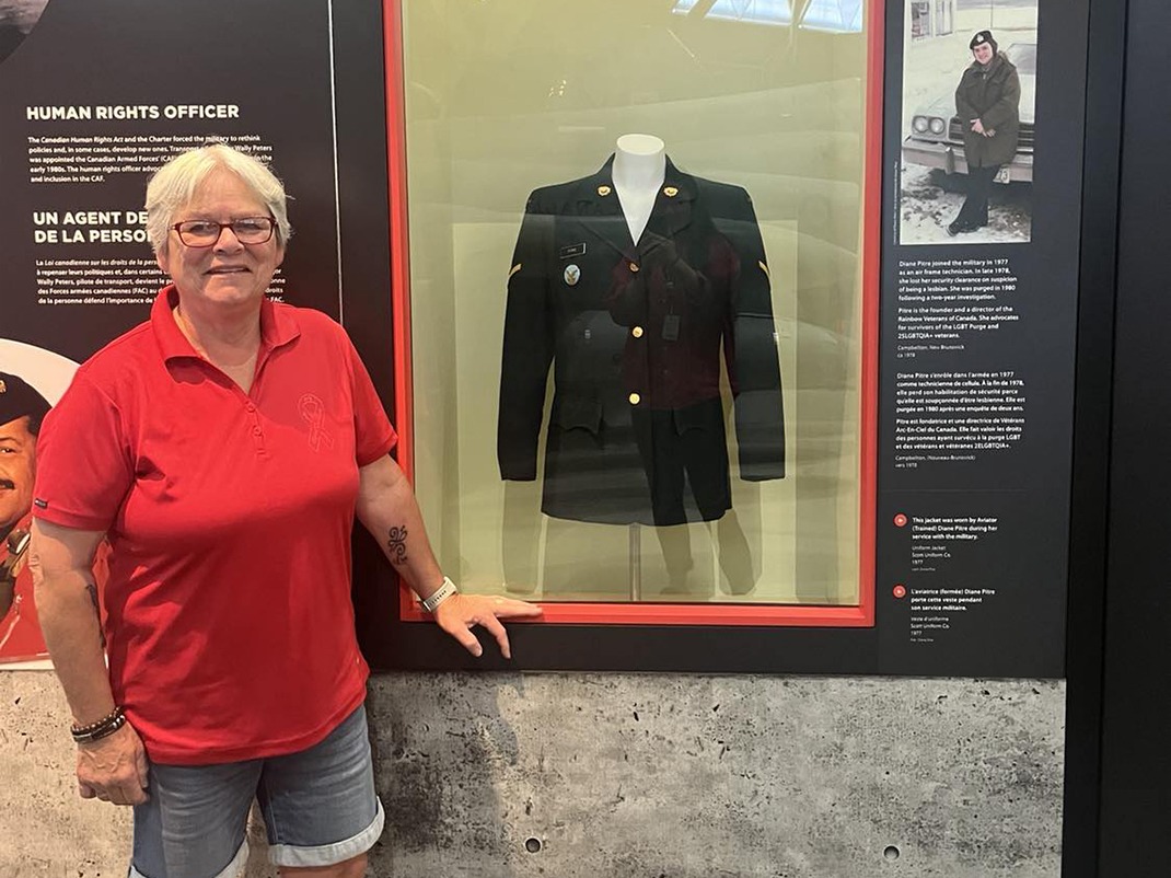 A person with short white hair wearing a red polo shirt and jean shorts, is standing smiling next to an exhibition display case that has a military jacket inside. There are text panels on either side of the display with an old picture of a woman standing in front of a car.