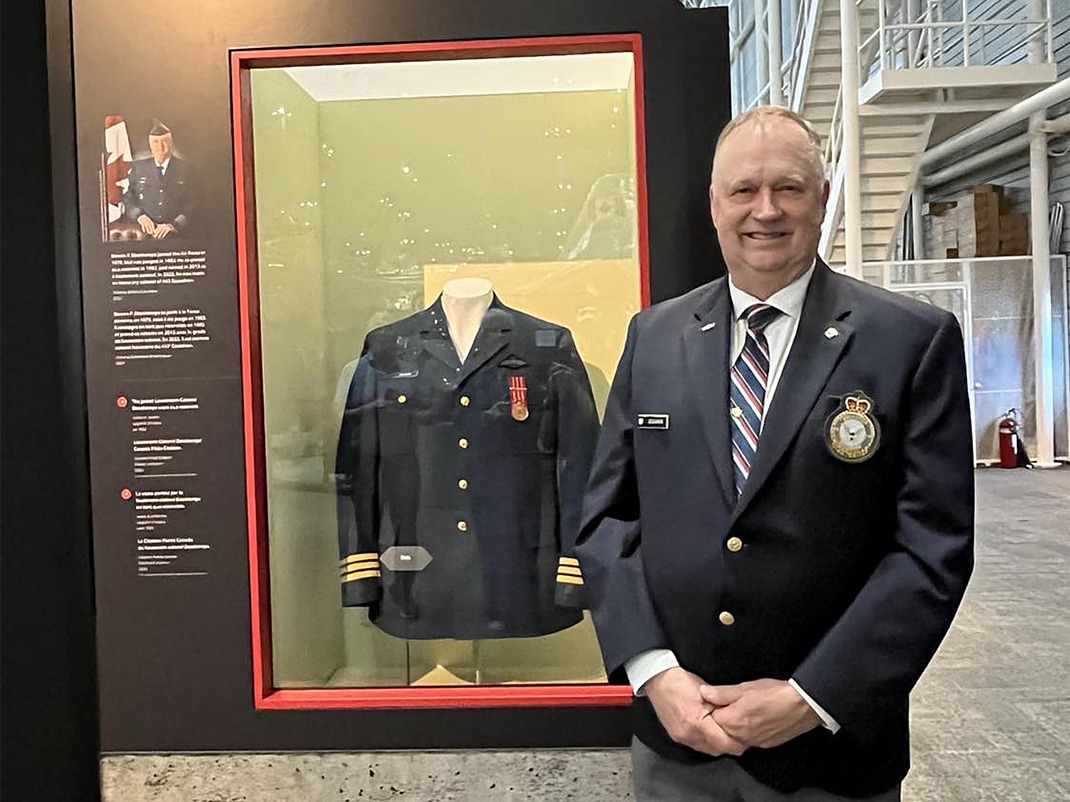 A man in a dark blazer with an embroidered crest and name tag stands smiling beside an exhibition display case containing a military uniform with medals. Next to the uniform is a photograph of the same man in full military dress and a text panel.
