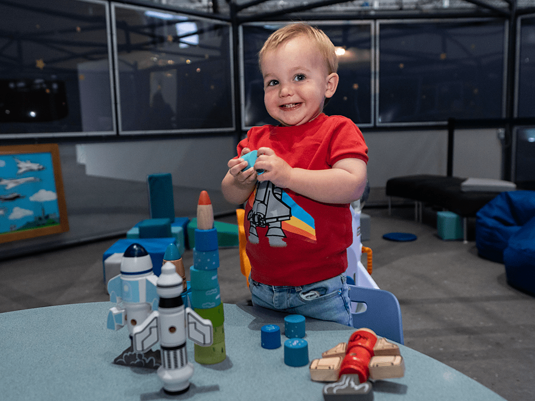 Un enfant en âge préscolaire aux cheveux blonds courts, vêtu d'un t-shirt rouge orné d'une fusée, sourit debout à côté d'une table sur laquelle sont disposés des jouets sur le thème de l'espace. À l'arrière-plan, on aperçoit des sièges confortables et d'autres jouets.