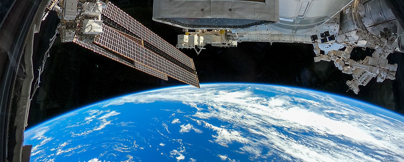 View of Earth’s blue surface and white clouds from space, with part of the International Space Station visible in the foreground, including solar panels and structural modules.