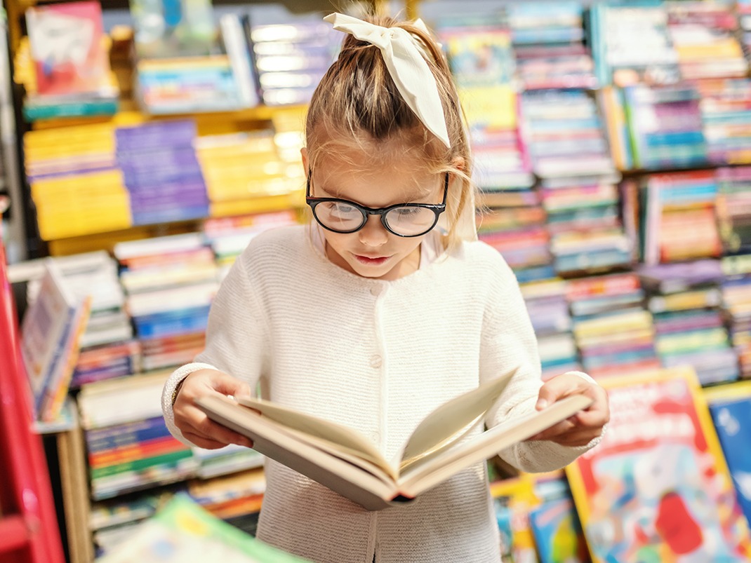 A child wearing a white sweater and a white hair bow stands in front of a densely packed bookshelf filled with colorful books. They are holding an open book and appear to be reading or examining it.