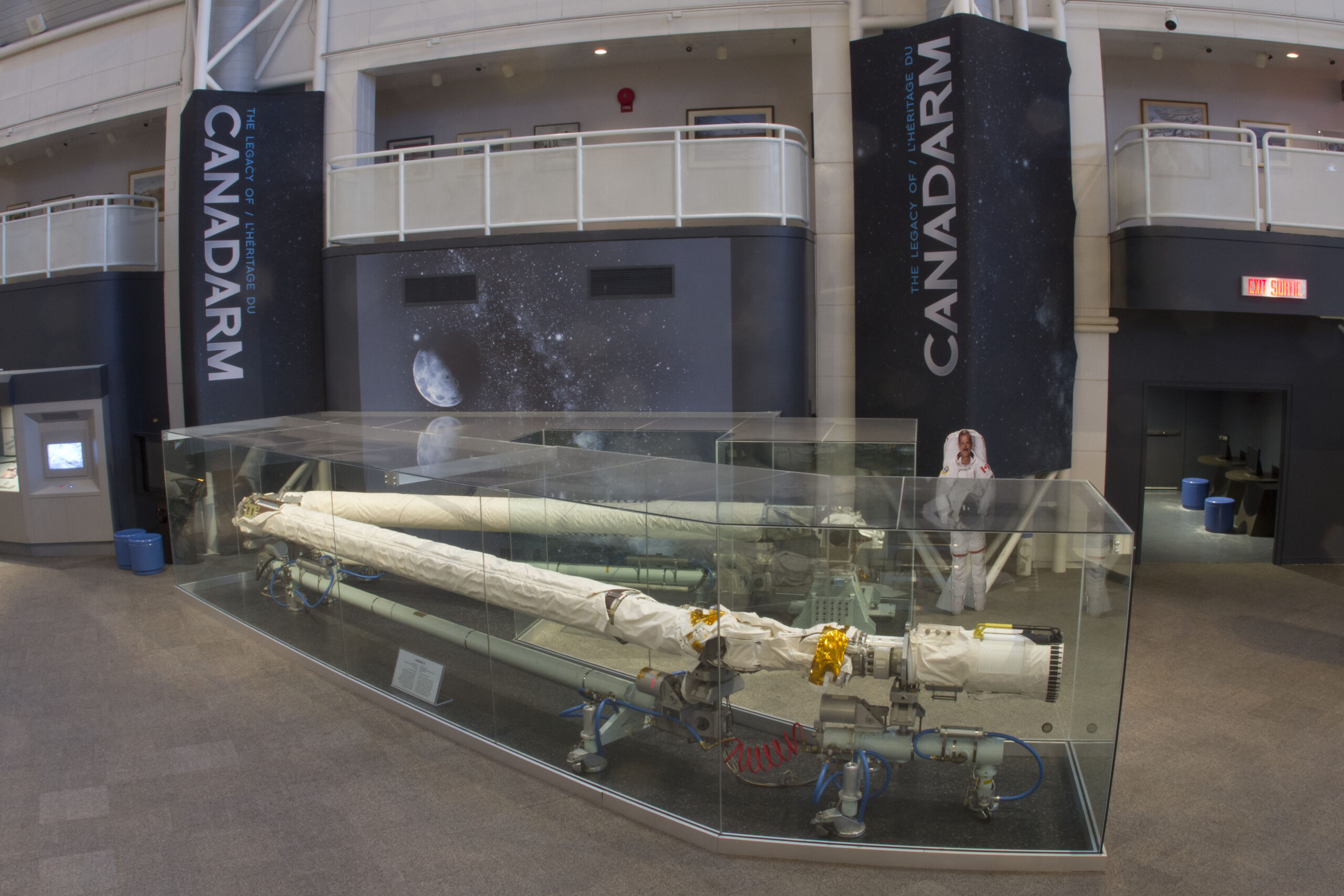 The Candarm inside of a display case on a museum floor. In the background there are two large pillars that say "Canadarm," and a balcony overhead with a white barrier.