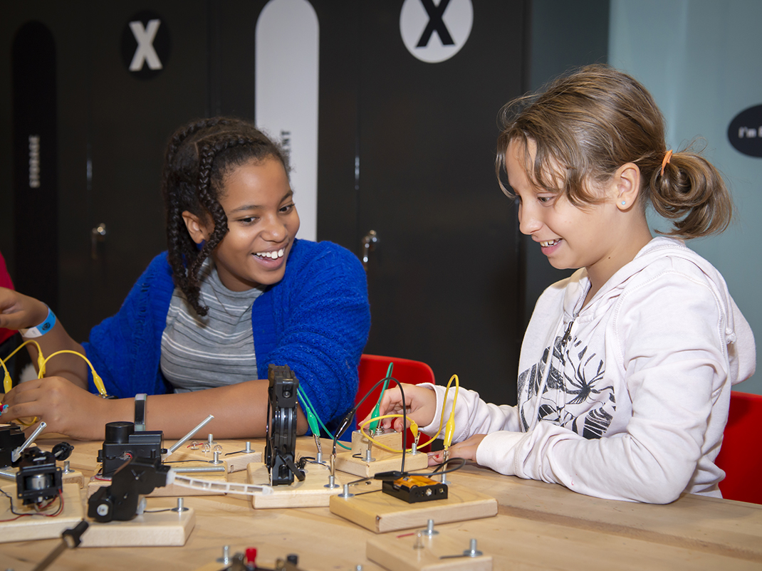 Two young girls are seated in red chairs at a wooden table, in front of them are homemade circuits made of little wood blocks, green and yellow wires and batteries. The girl on the left is smiling at the girl on the right as she tests out the circuit with a smile on her face.