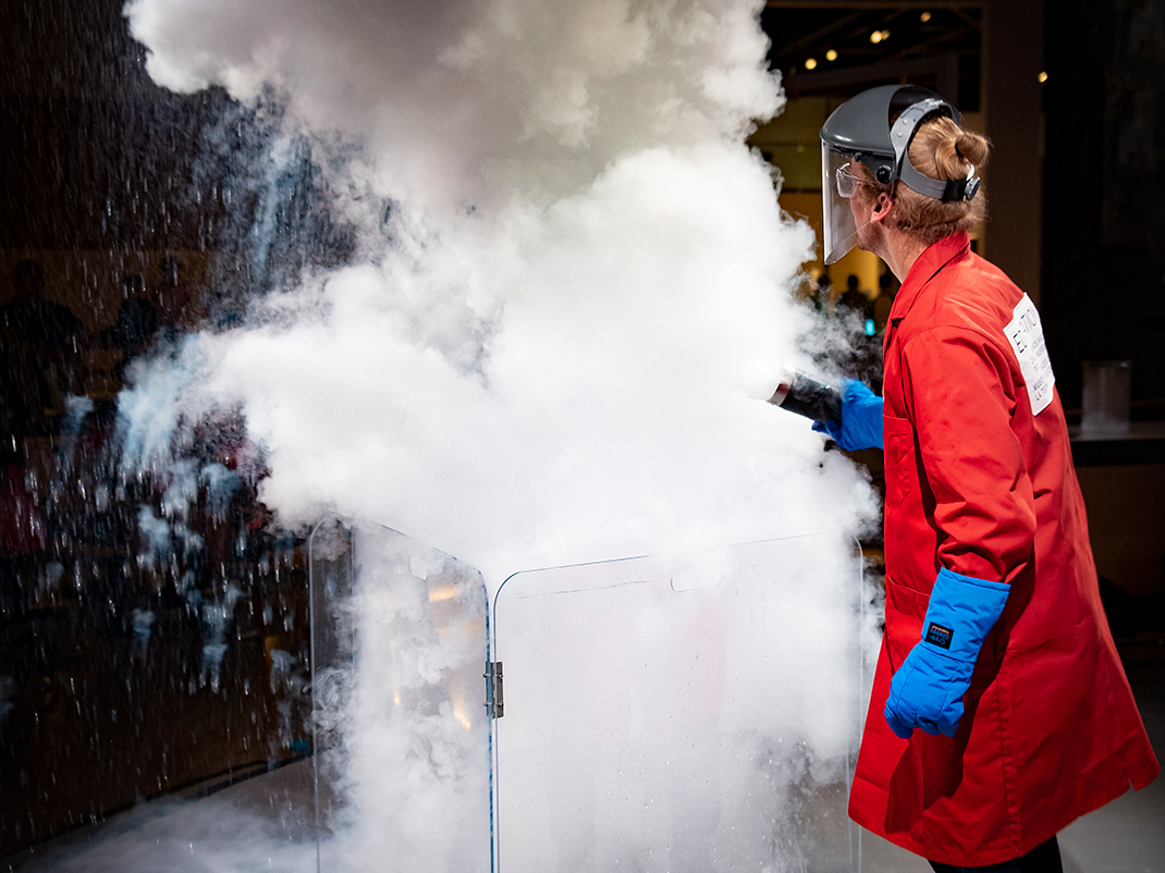 A museum guide on the demonstration stage is dressed in a red lab coat, blue protective gloves, and a protective visor. He holds a canister as he steps backward from an experiment he has just executed, which has produced a large plume of white powder and smoke.