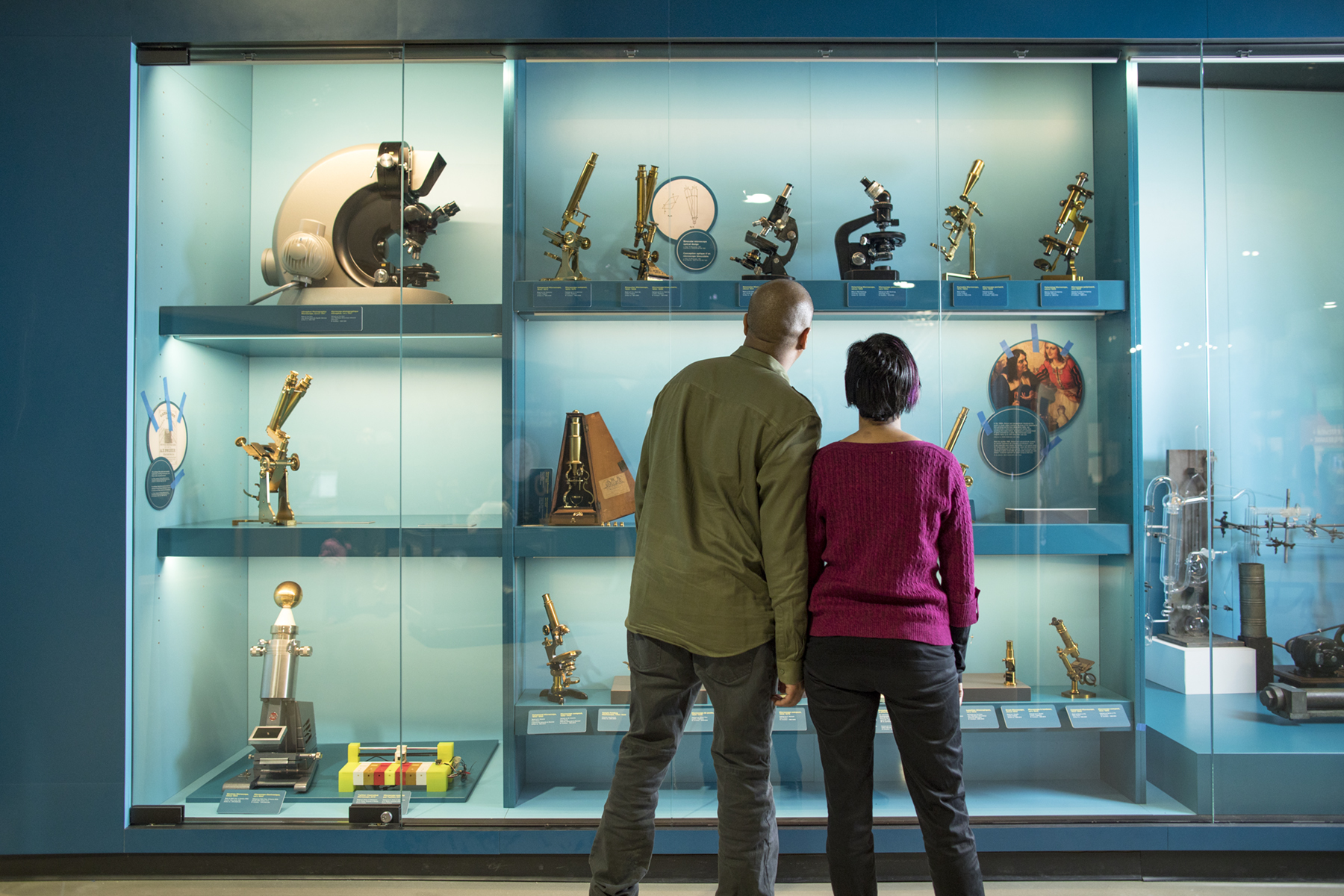 Two museum visitors stand side by side, admiring a brightly lit display case filled with a variety of historical microscopes and related scientific instruments. The case is organized in rows, showcasing items ranging from antique brass microscopes to more modern designs.