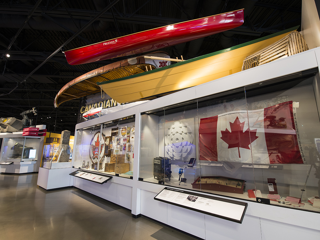 Museum exhibit displaying Canadian flags, canoes, and historical artifacts in a glass case.