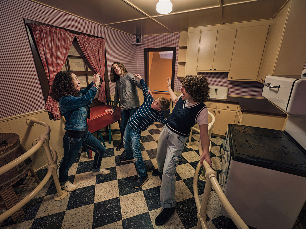 A mom and three kids of different ages are standing in a slanted kitchen with a black and white checkered floor. The mom is taking a picture of the kids as they lean to one side with one of their hands up in the air.