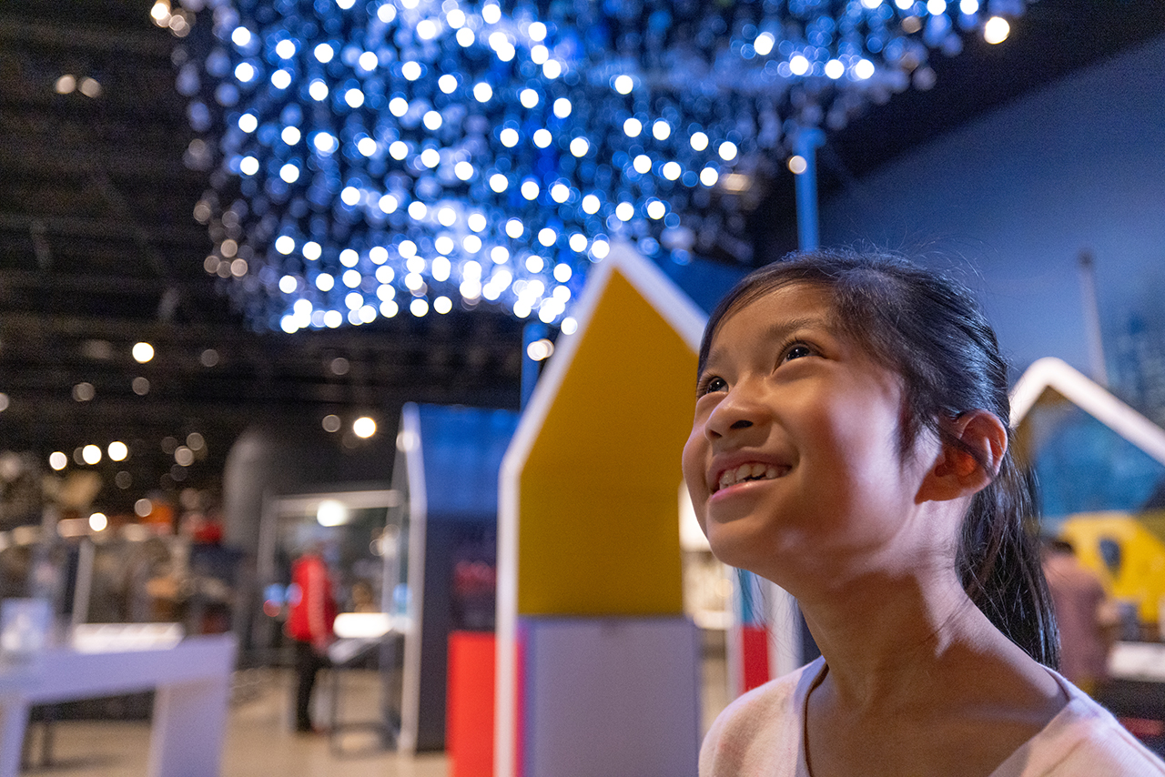 A young child smiles, looking up at an installation of lights within a museum. In the background there are colourful house-shaped exhibition panels.