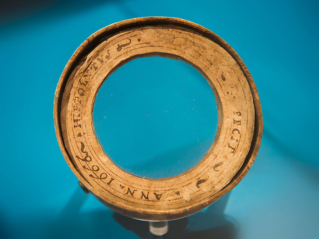 A close-up of an antique lens or optical device with inscriptions around the edge, set against a blue background.