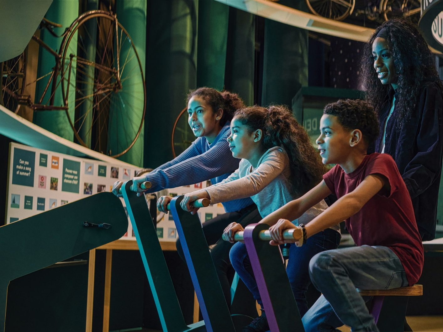 Three children ride stationary bikes indoors at the museum, with an adult supervising. Colorful backdrop with bicycle wheels and educational displays in the background.