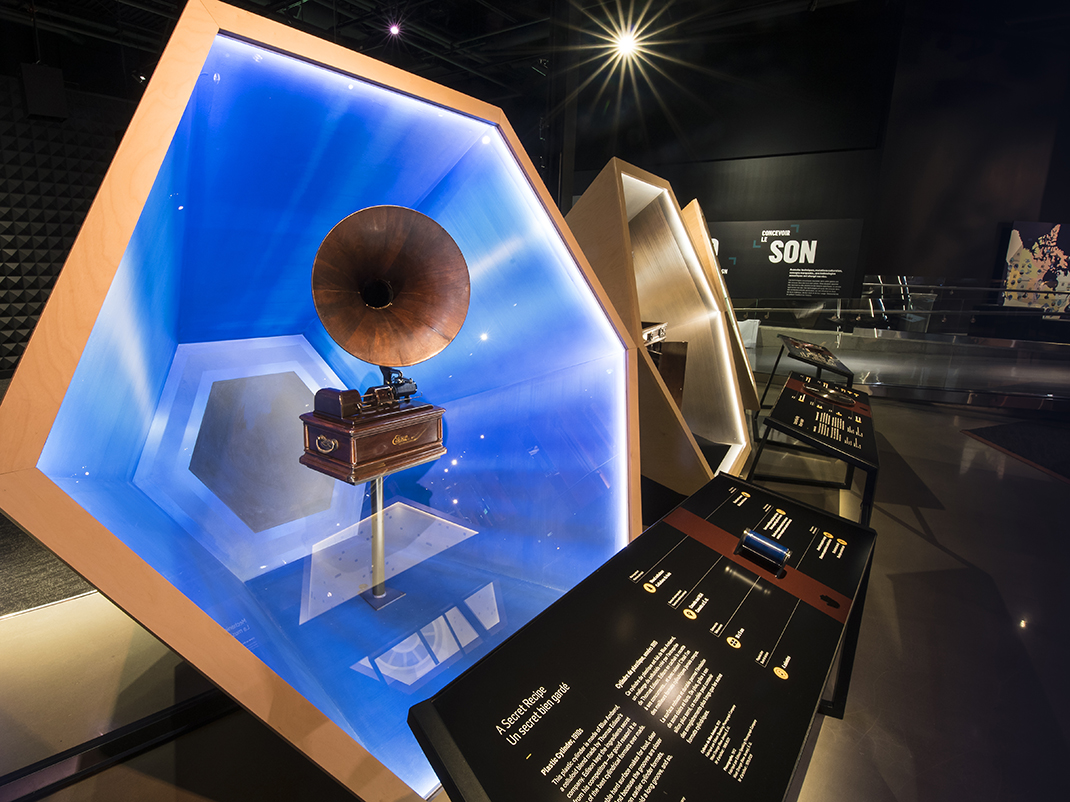 An antique gramophone is on display in a blue hexagon display case. Museum panels of text are in the foreground.